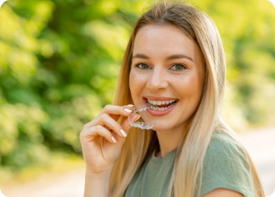 Smiling blonde woman holding a clear aligner outdoors